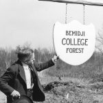 A man reaching out, touching, and looking at a sign for the Bemidji College Forrest in 1947