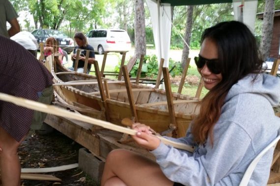 A female student holding a piece of wood beside a large wooden canoe
