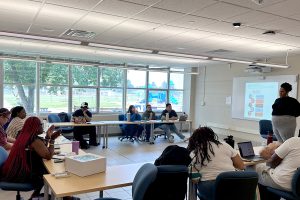A woman gives a presentation to a group of students seated at tables. A PowerPoint presentation is on a screen behind her.
