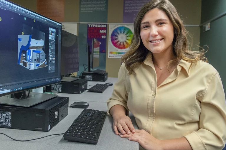 a student smiling and sitting at a computer with 3d graphics on it