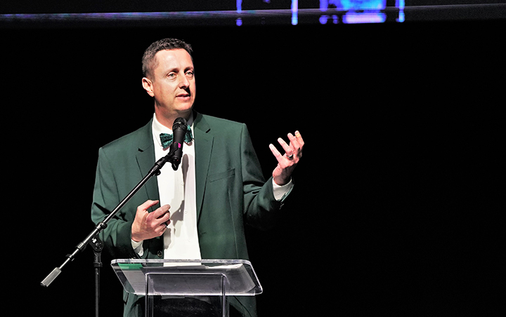 President John Hoffman wearing a green suit jacket and bowtie giving a speech at a podium