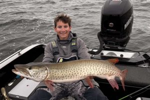 BSU grad student Baylor Short sits at the back of a boat with a large Mercury outboard motor behind him. He is smiling and holding a large muskie in front of him with both hands.