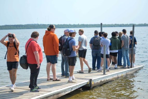 Students from the Engineering Problem Solving course watch their classmates paddle their boats on Lake Bemidji