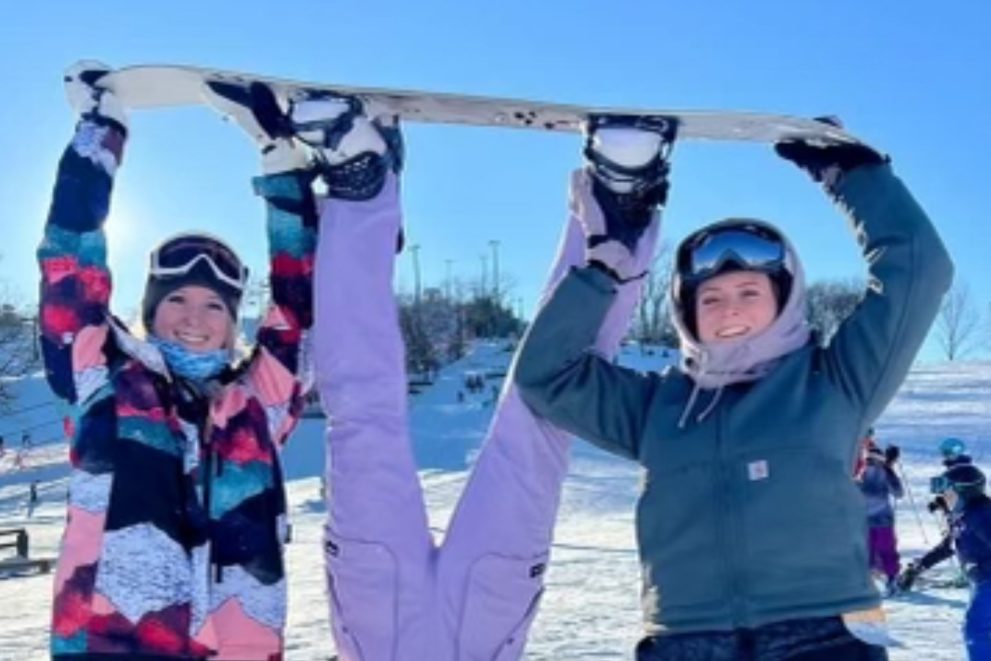 Two BSU college students in snowboarding gear holding a third student upside down by her board