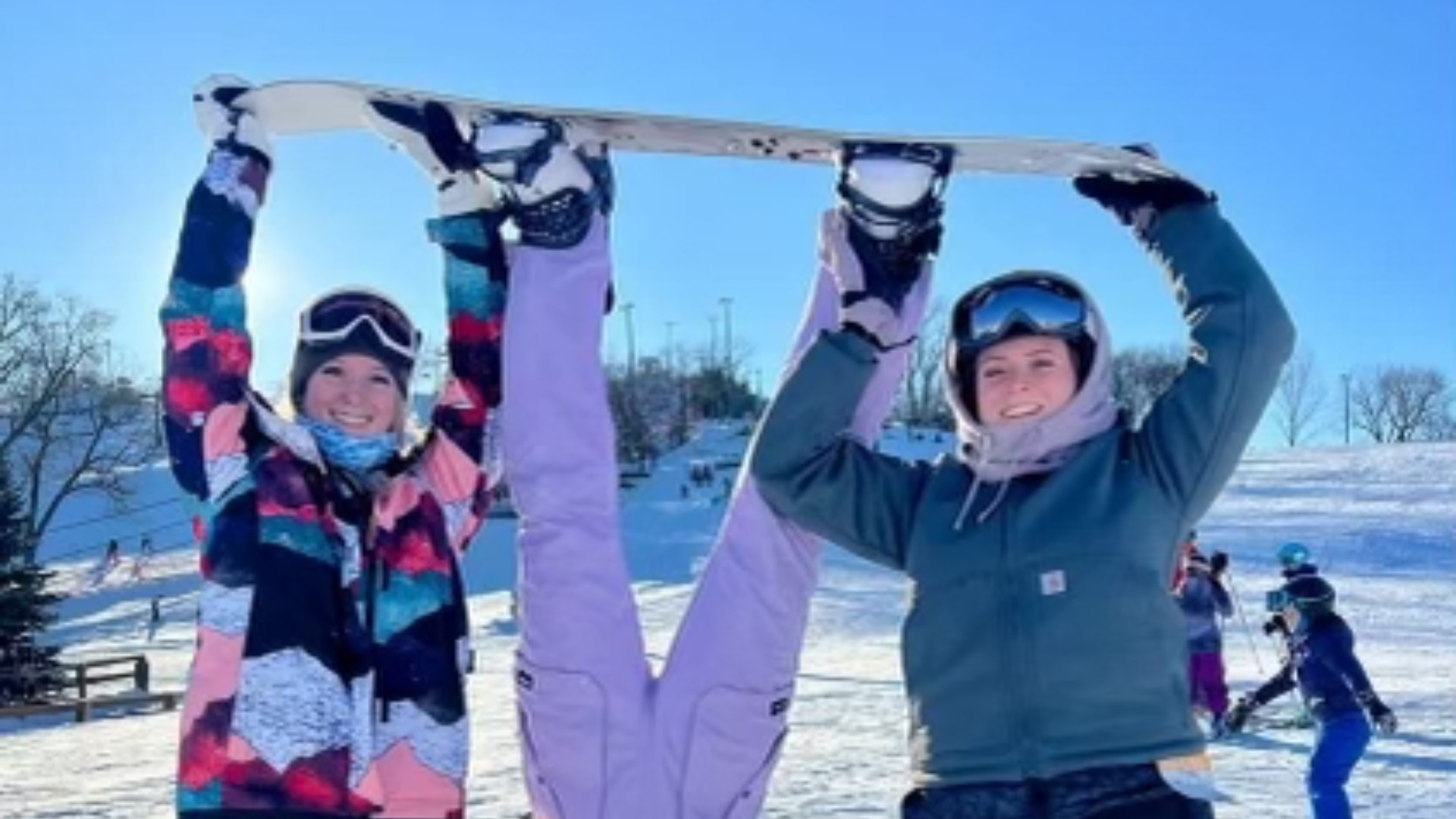 Two BSU college students in snowboarding gear holding a third student upside down by her board