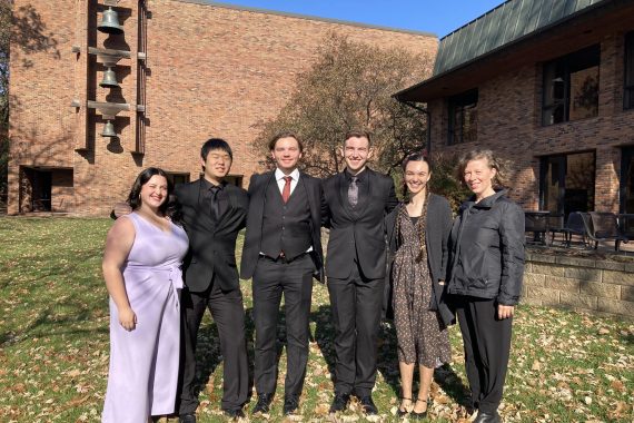Five BSU students pose outdoors in front of a brick building with their accompanist.