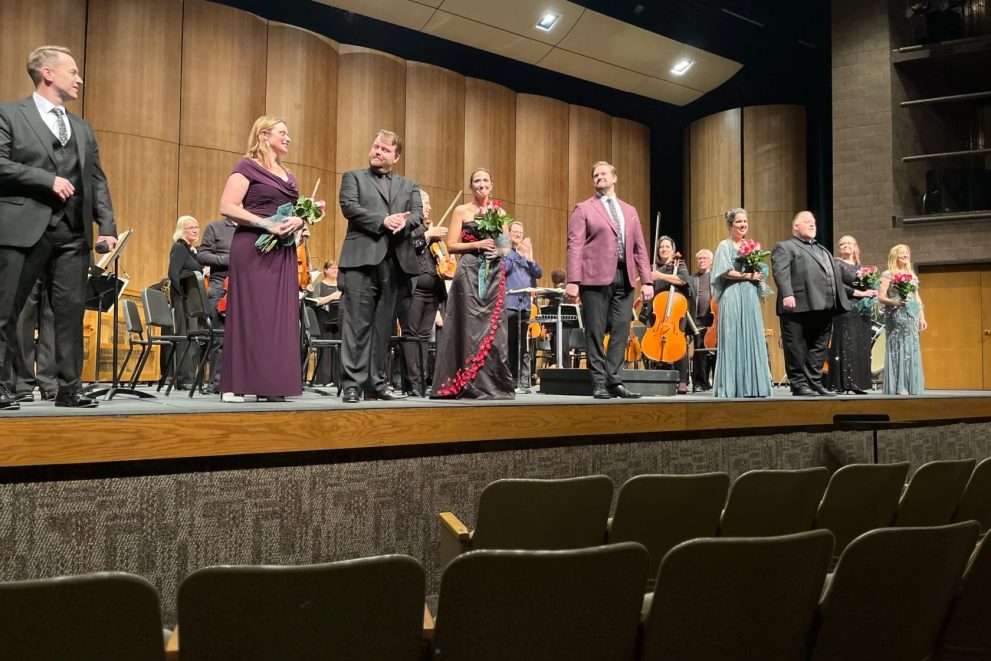 A photo of 9 singers in a line on stage reacting to audience applause at the October 28 Fargo Moorhead Opera concert