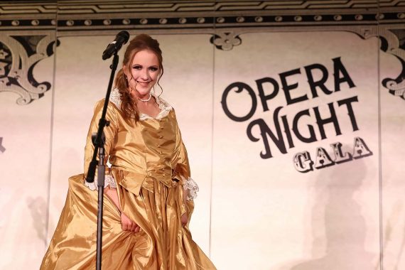 A woman with dark blonde hair in a gold dress performs in front of a light-colored backdrop which reads "Opera Night Gala"