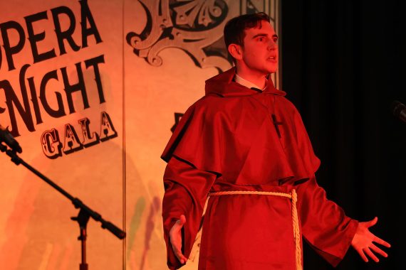 A man wearing religious robes performs in front of a backdrop which reads "Opera Night Gala". The stage is bathed in red light and has an ominous feeling.