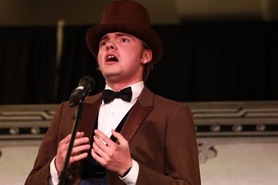 A man in a brown suit with a bow tie and top hat sings in front of a backdrop which reads "Opera Night Gala"