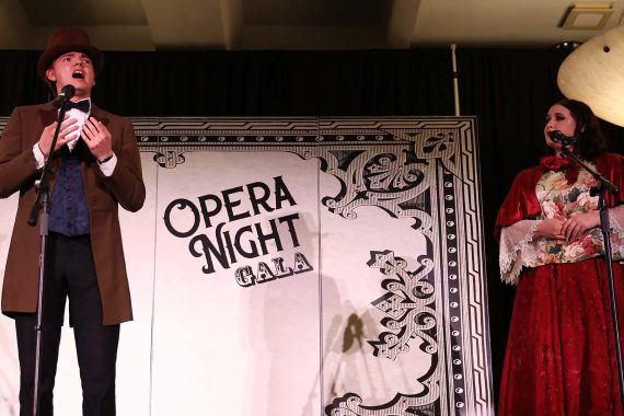 A man in a brown suit with a bow tie and top hat sings with a woman in a red dress with a red shawl in front of a backdrop which reads "Opera Night Gala"