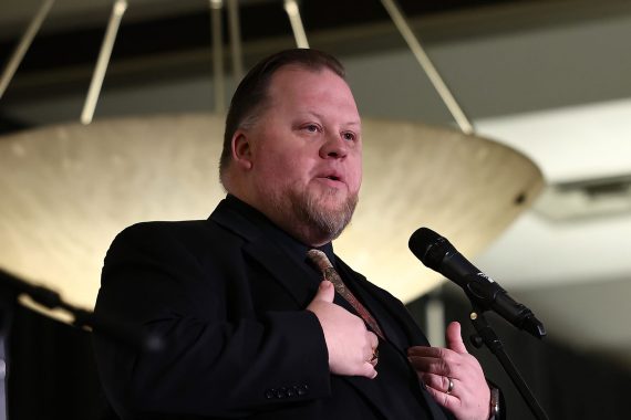 A man speaks into a microphone, gesturing with his hands in front of him.