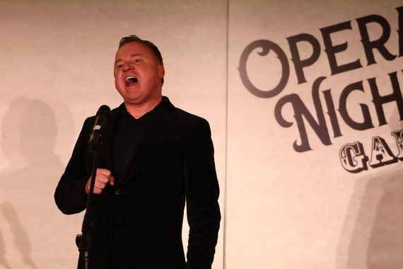 A man in a dark suit sings with his right hand clenched in front of him in front of a banner that says "Opera Night Gala"