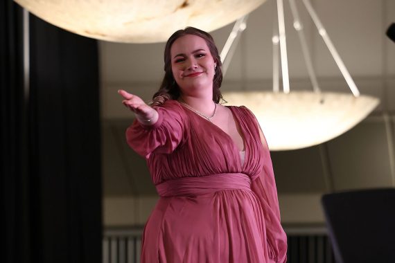 A woman in a pink dress gestures toward the camera, acknowledging her accompanist after her performance.