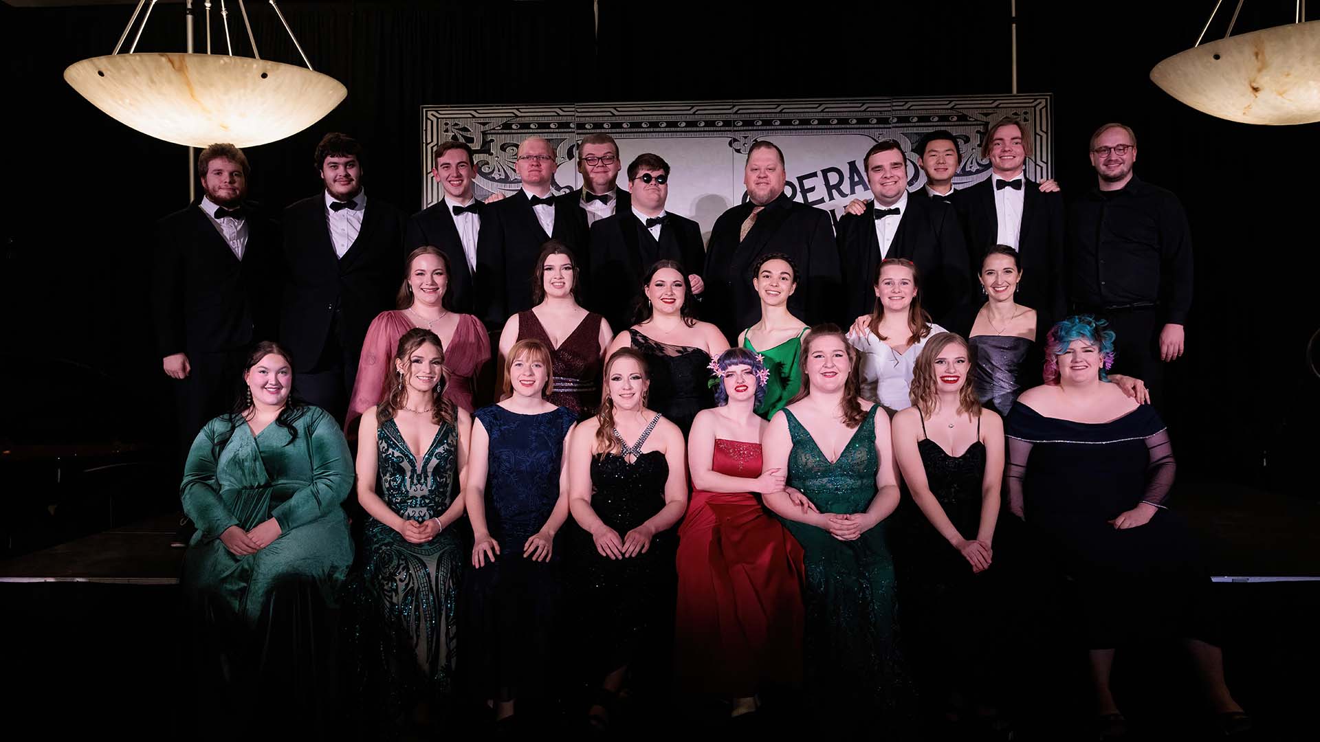 A group of 25 performers wearing formal attire, posed in three rows.