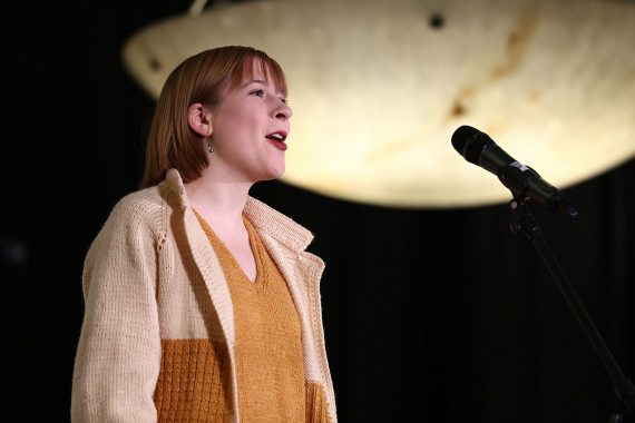 A woman in a tan, brown and mustard colored knit outfit sings in front of a light.