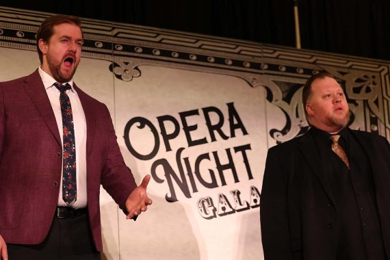 A man with a beard in a brown jacked and colorful tie sings a duet with a man wearing a dark suit. Behind them is a banner which reads "Opera Night Gala"