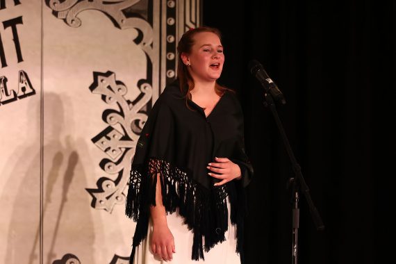 A woman with a black shawl and white dress is singing.