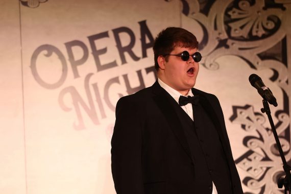 A man wearing dark, round sunglasses and a tuxedo sings in front of a banner which reads "Opera Night Gala"