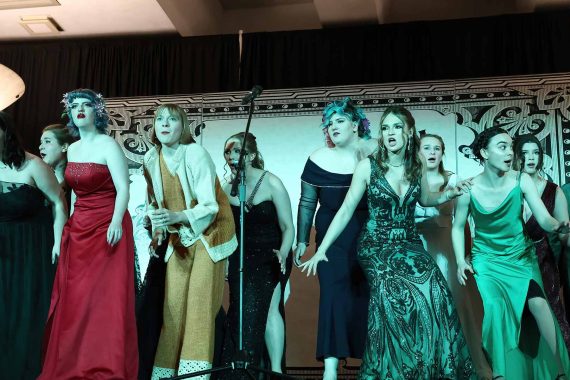 A chorus of 11 women are singing on a stage. They're looking very serious.