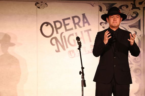 A man in a black suit and a black Fedora with a a small red feather on the left side sings in front of a banner which reads "Opera Night Gala"