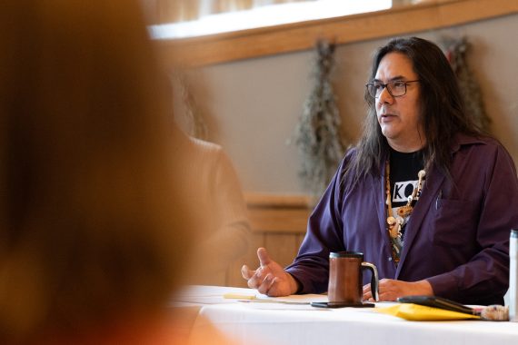 A man with long hair and glasses, and wearing a purple long-sleeved shirt, speaks at a table.