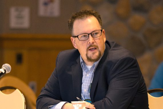 A man with glasses and a goatee, wearing a blue jacket and a blue plaid shirt, speaks with his hands folded on a table in front of him