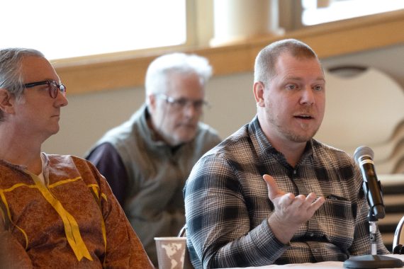 A man wearing a plaid shirt is speaking at a table. His right hand is out in front of him, with his palm raised