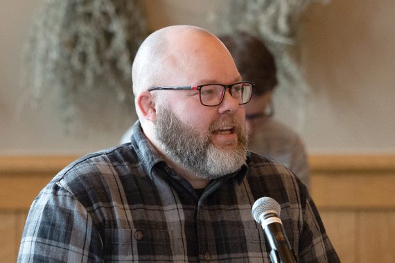 A man with a beard and glasses, wearing a plaid shirt, speaks into a microphone
