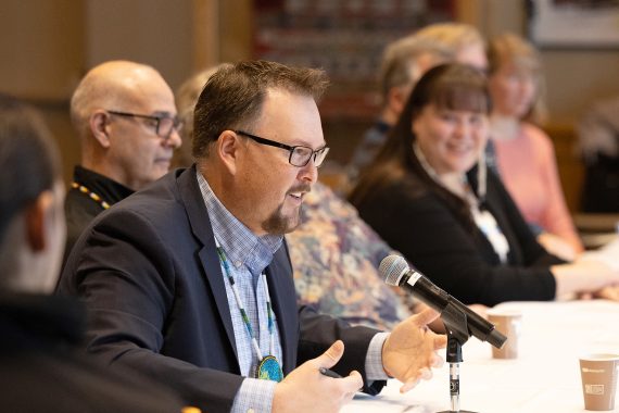 A man with glasses and a goatee, wearing a blue jacket and a blue plaid shirt, speaks with his hands folded on a table in front of him