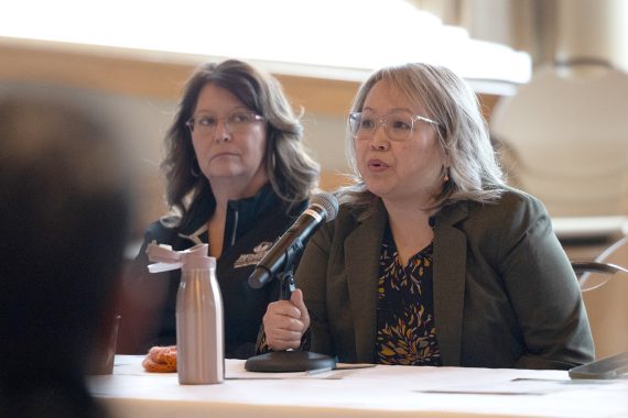 A woman with glasses wearing a brown suit speaks into a microphone