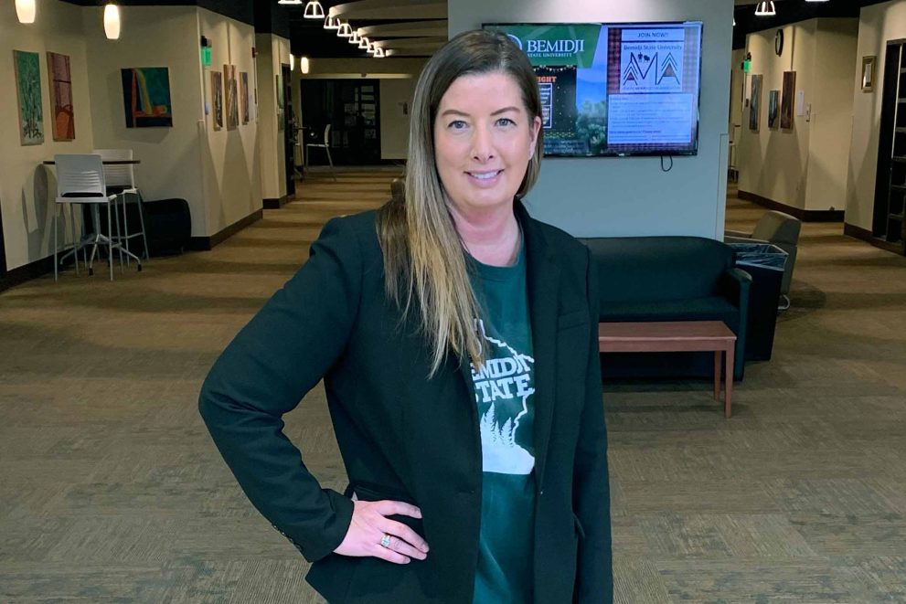 Kelly LaVenture is wearing a black jacket over a green BSU t-shirt. She is standing in the basement of Memorial Hall in front of a television.