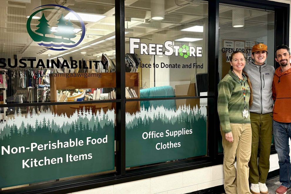 Three people standing in front of windows decorated with decals of pine trees. A logo on the window says "Free Store" in English and Ojibwe.