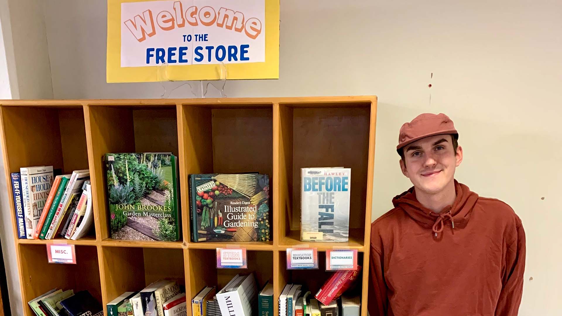 A male student in a red shirt and orange jacket poses by a bookcase filled with items in BSU's free store. A sign above the bookcase reads "Welcome to the Free Store"