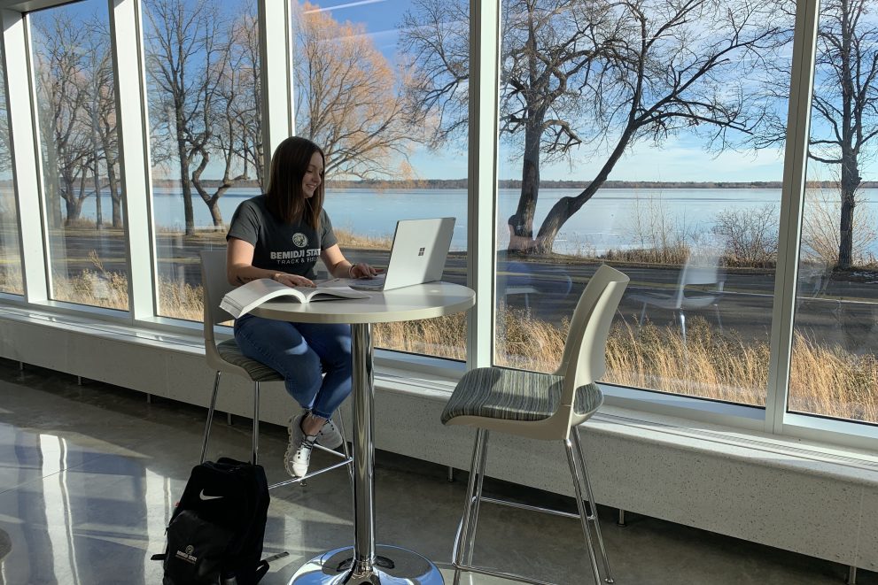 Olivia Olson sitting with her laptop in a building with Lake Bemidji in the background