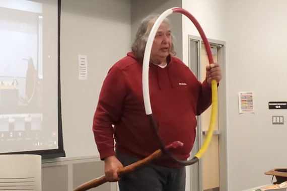 A man with long, grey hair and wearing a red long-sleeved shirt holds a painted snow snake in his right hand and a hoop with quarters painted white, red, yellow and black in his left.
