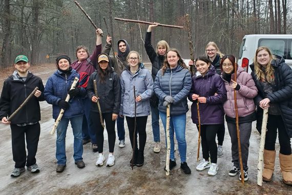 A group of students are posing outdoors with sticks they will use to make snow snakes.