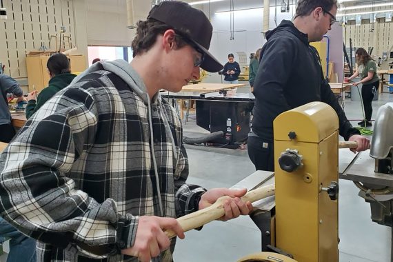 A student in a black and white plaid jacket and a black baseball cap is using a yellow machine to prepare a stick for painting as a snow snake.