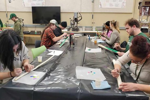 A group of students are painting sticks on a long table covered in black plastic.