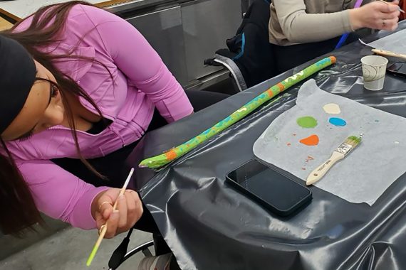 A student is painting sticks on a long table covered in black plastic.