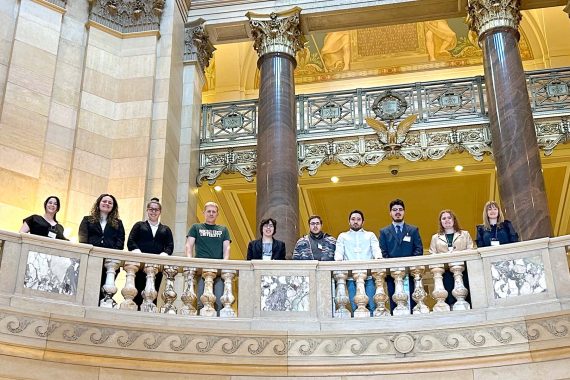 A group photo of BSU's Posters at the Capitol students and faculty, posed by a stone rail. The photo is shot from below; the group is on a higher level looking down