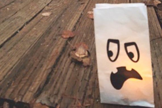 A white paper sack votive, lit from the inside, has a spooky ghost face on it. It sits on a boardwalk.