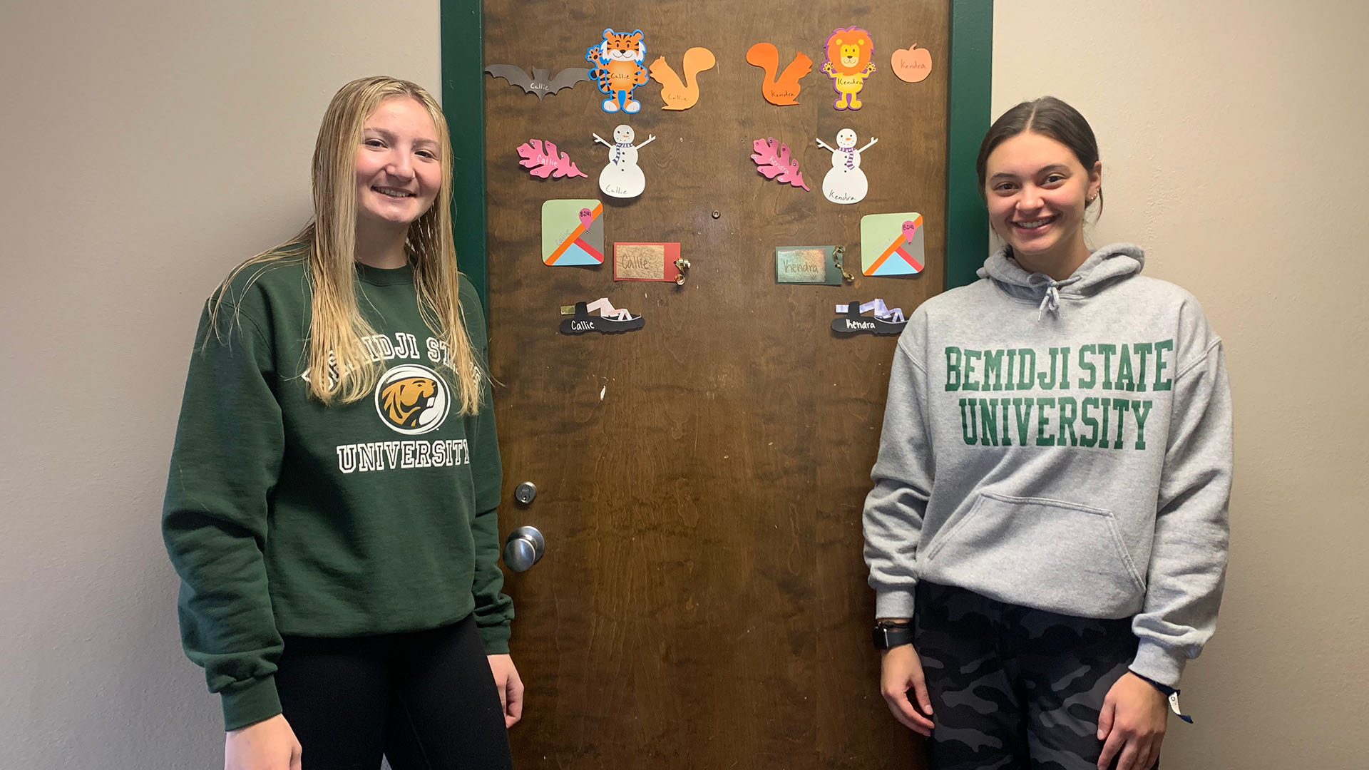 Two students wearing BSU sweatshirts standing in front of a residence hall door that is decorated with more than a dozen cutout paper nametags.