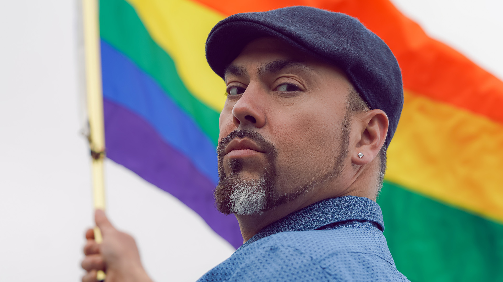 An Indigenous man in a blue shirt with a beard and a blue scally cap waves a pride flag