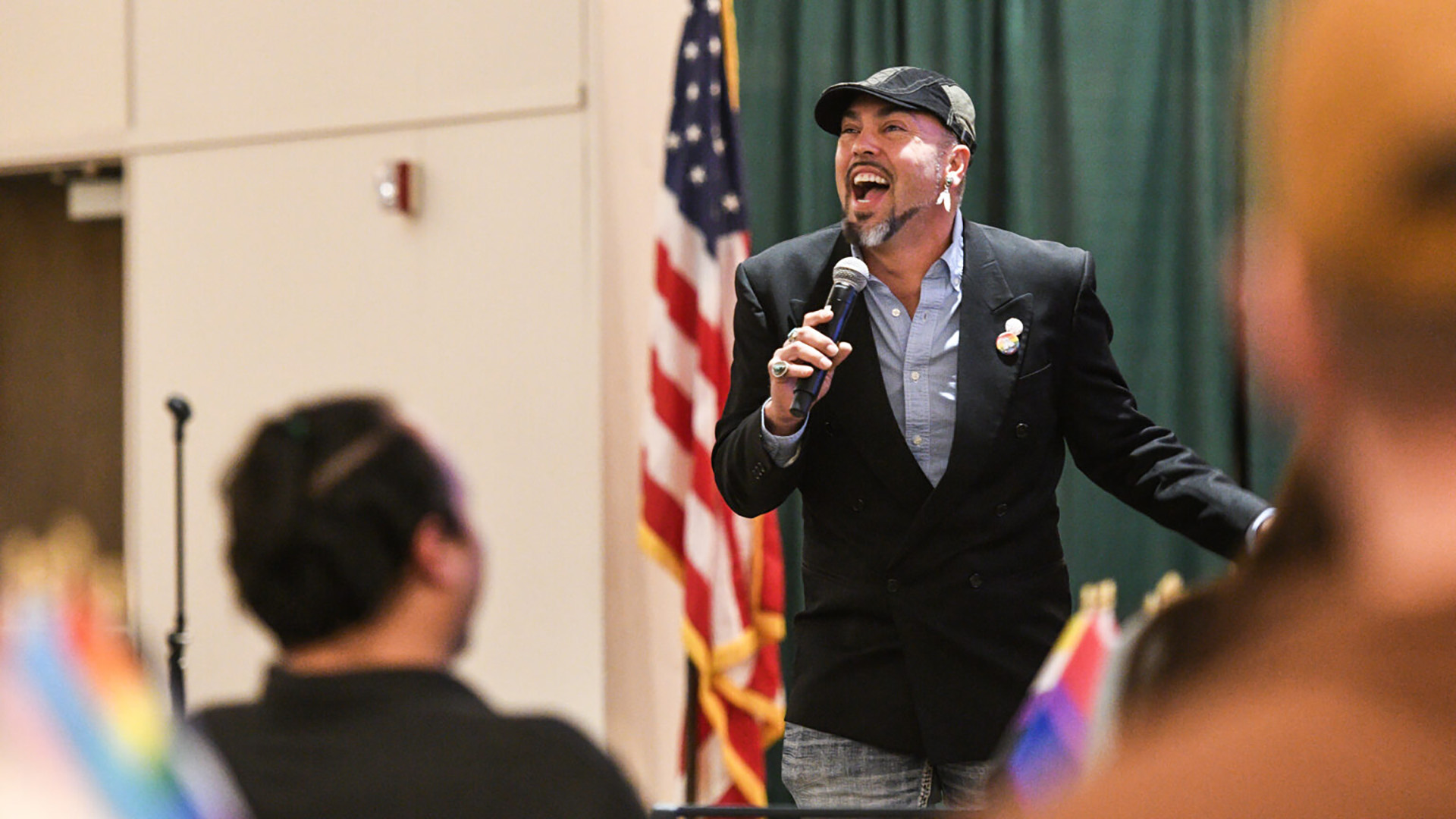 A man wearing a dark suit jacket and jeans with a dark blue and grey scally cap speaks into a microphone