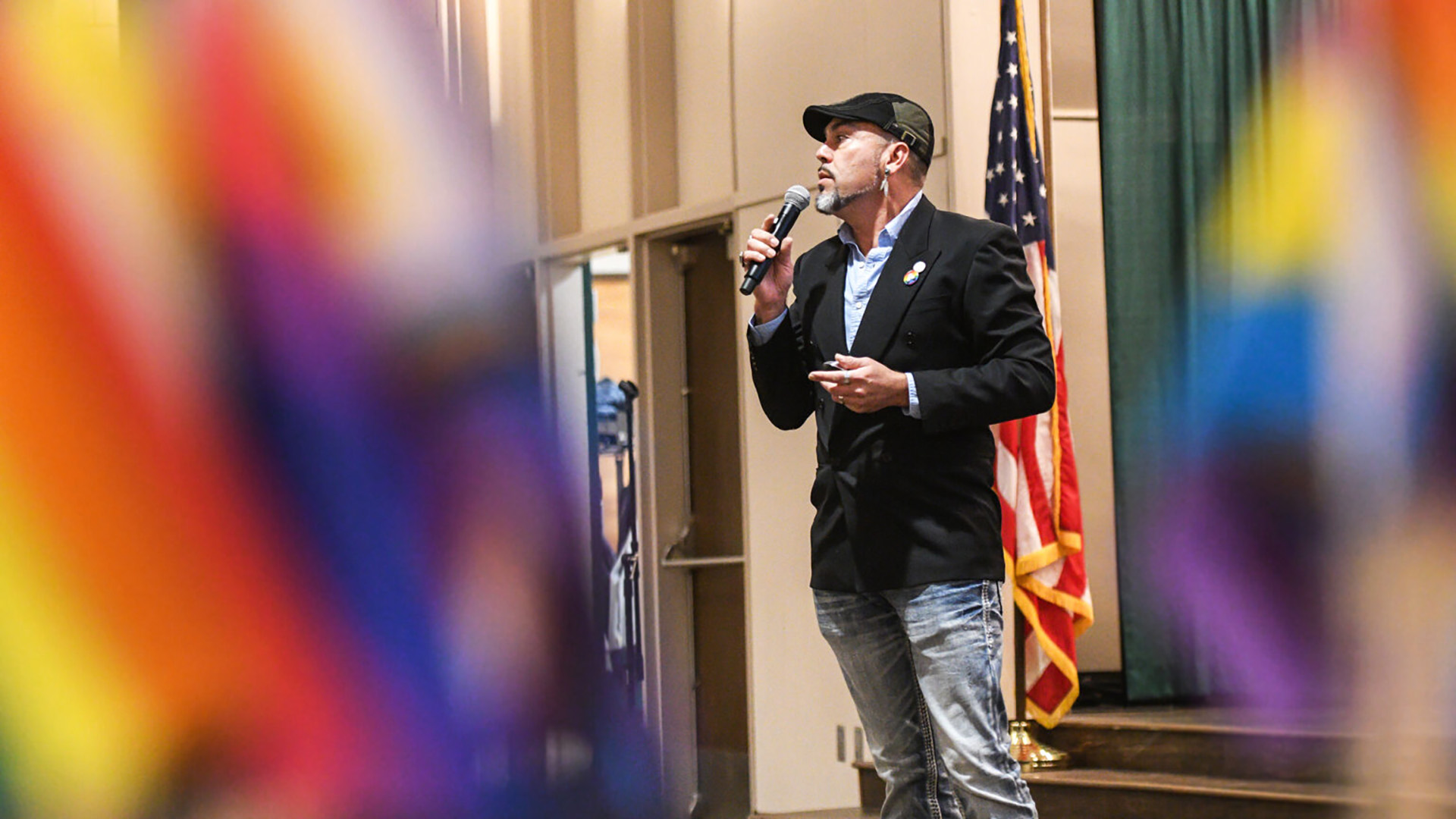 A man wearing a dark suit jacket and jeans with a dark blue and grey scally cap speaks into a microphone