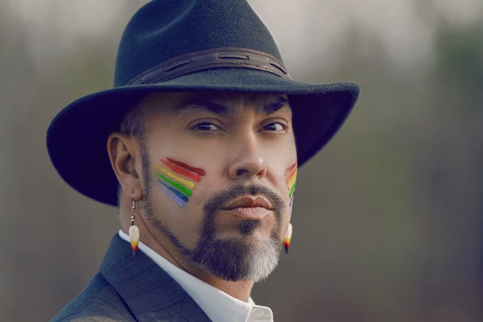 A man with a goatee wearing a black fedora, a dark suit and white shirt. He has rainbow paint on each cheek.