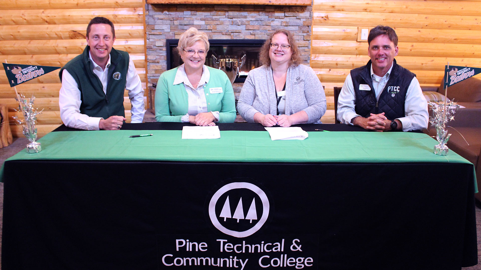  Two administrators from BSU and two from Pine Technical & Community College sit at a green table with Pine Technical College's logo on the front. There are papers and pens on the table for contract signings.