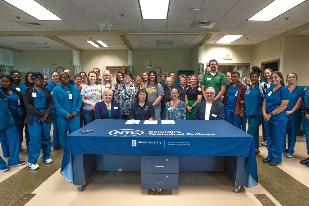 NTC nursing students stand behind a table draped with a blue table cloth and the NTC logo. BSU and NTC administrators sit at the table and are about to sign contracts.