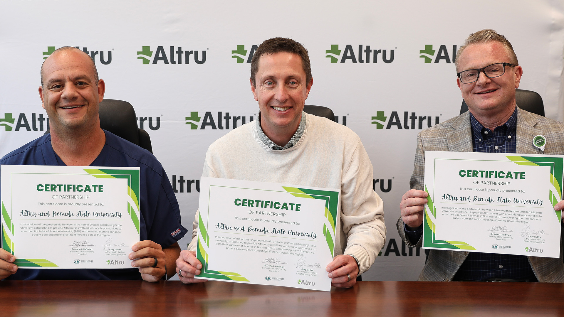 Three people sit in front of a backdrop with Altru Health logos on it. They are holding signed certificates that symbolize the partnership between BSU and Altru Health.
